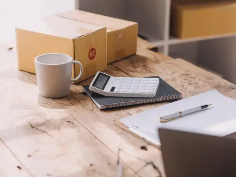 A calculator on a notebook, a clipboard with a pen and a mug beside taped cardboard boxes on a wooden table