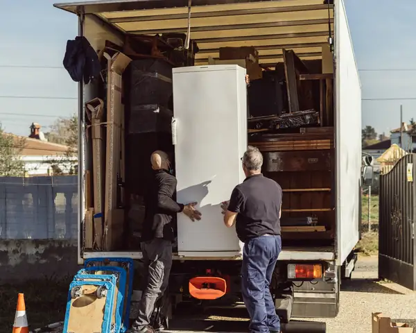 Two movers seen from behind lift a white refrigerator into the open back of a box truck loaded with wrapped furniture, in bright sunshine