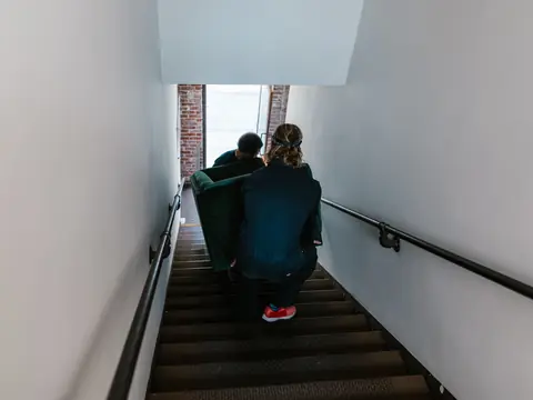 Two movers seen from behind carry a green sofa down a narrow stairway toward an open street door